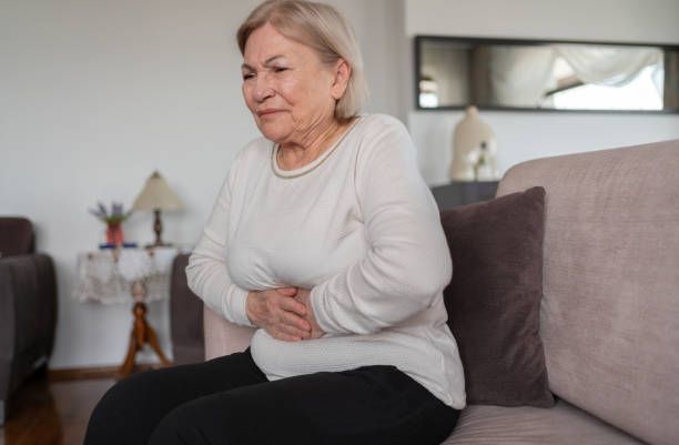 An older woman sitting on a couch holds her stomach with both hands and appears to be experiencing abdominal discomfort.