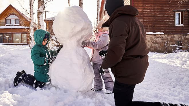 Family building a snowman together in a snowy yard near wooden houses.