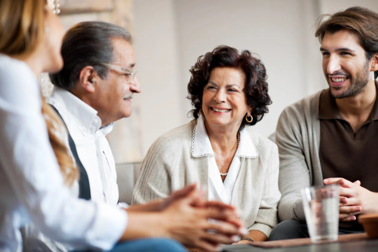 Four family members chat and smile in a bright room, wearing white, beige, and brown clothing with soft knit textures.