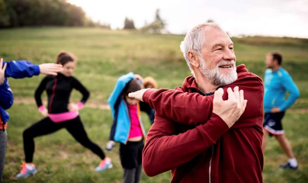 Senior man stretching his shoulders during an outdoor group exercise session in a park.