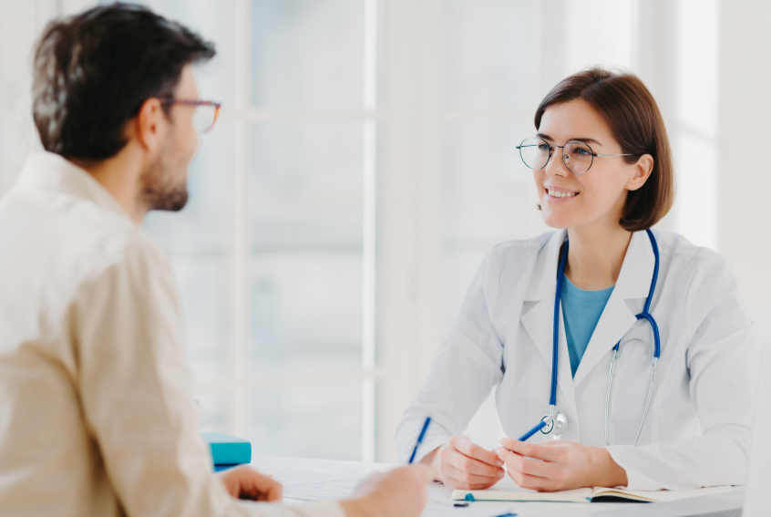 Female doctor in a white coat with a blue stethoscope smiles while speaking with a patient in a bright, softly lit office.